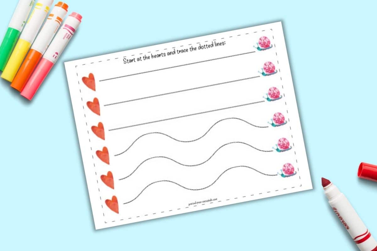 A top down flatlay mockup of a printable fine motor prewriting practice worksheet. On the left is a line of five hearts next to three straight lines and three wavy dotted lines. The lines lead to a Valentine's snail. The page is on a blue background with colorful child's markers.