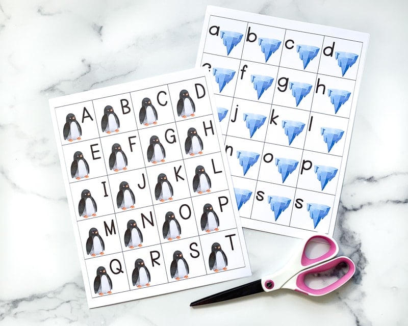 A preview of two free printable alphabet matching cards printable pages. Each page has 20 squares with the letters a-t. On the left is a page with a penguin on each card and uppercase letters. On the right is a page with an iceberg on each card and lowercase letters. A pair of scissors is on a marble surface near the cars.