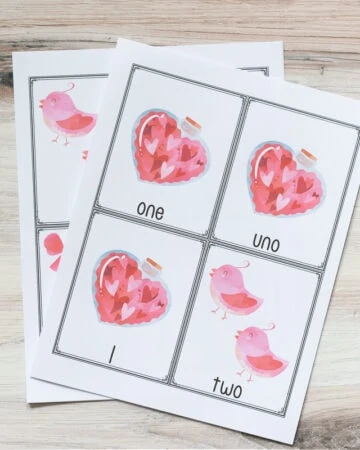 two free printable pages of Valentine's themed counting cards for preschoolers. The top page has three cards for the number 1 with a bottle of hearts and one card for two with two pink birds. The cards are in English, Spanish, and a numeral