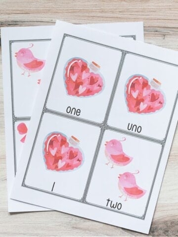 two free printable pages of Valentine's themed counting cards for preschoolers. The top page has three cards for the number 1 with a bottle of hearts and one card for two with two pink birds. The cards are in English, Spanish, and a numeral