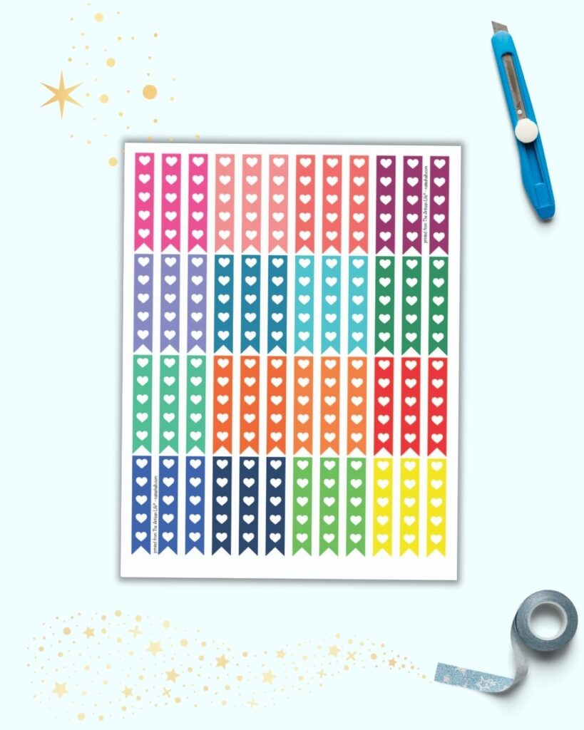 A sheet of printable stickers on a light blue background. The stickers are narrow vertical flags with 5 hearts to use as checkboxes. There are 16 different rainbow colors with three flags each per color. The sheet is on a light blue background with blue washi tape and a blue hobby knife.