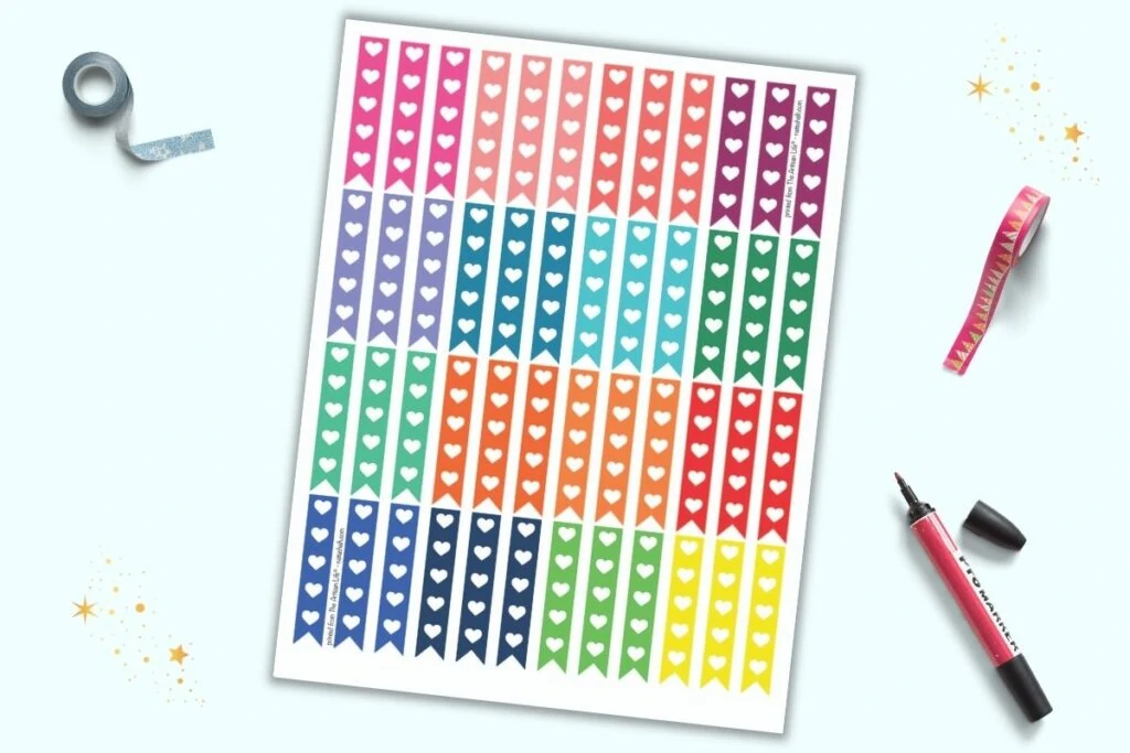 A sheet of printable stickers on a light blue background. The stickers are narrow vertical flags with 5 hearts to use as checkboxes. There are 16 different rainbow colors with three flags each per color. The sheet is on a light blue background with blue washi tape, red washi tape, and a red marker.