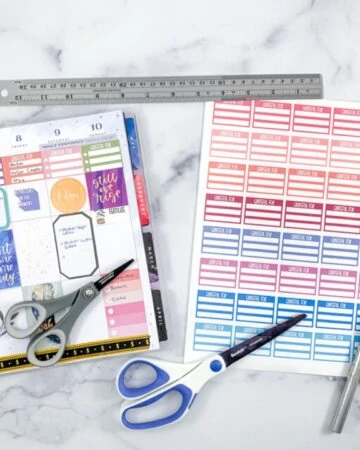 An open Happy Planner next to a page of colorful printed planner stickers. There are also two pairs of scissors, a metal ruler, and a metal hobby knife.