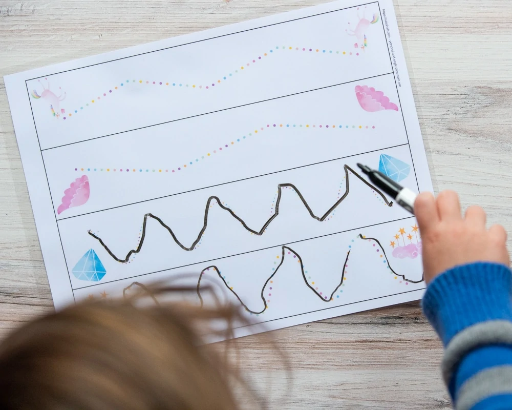 An overhead view of a young child's hand with a black marker tracing the lines on a unicorn themed prewriting practice page. The page is on a wood surface. 