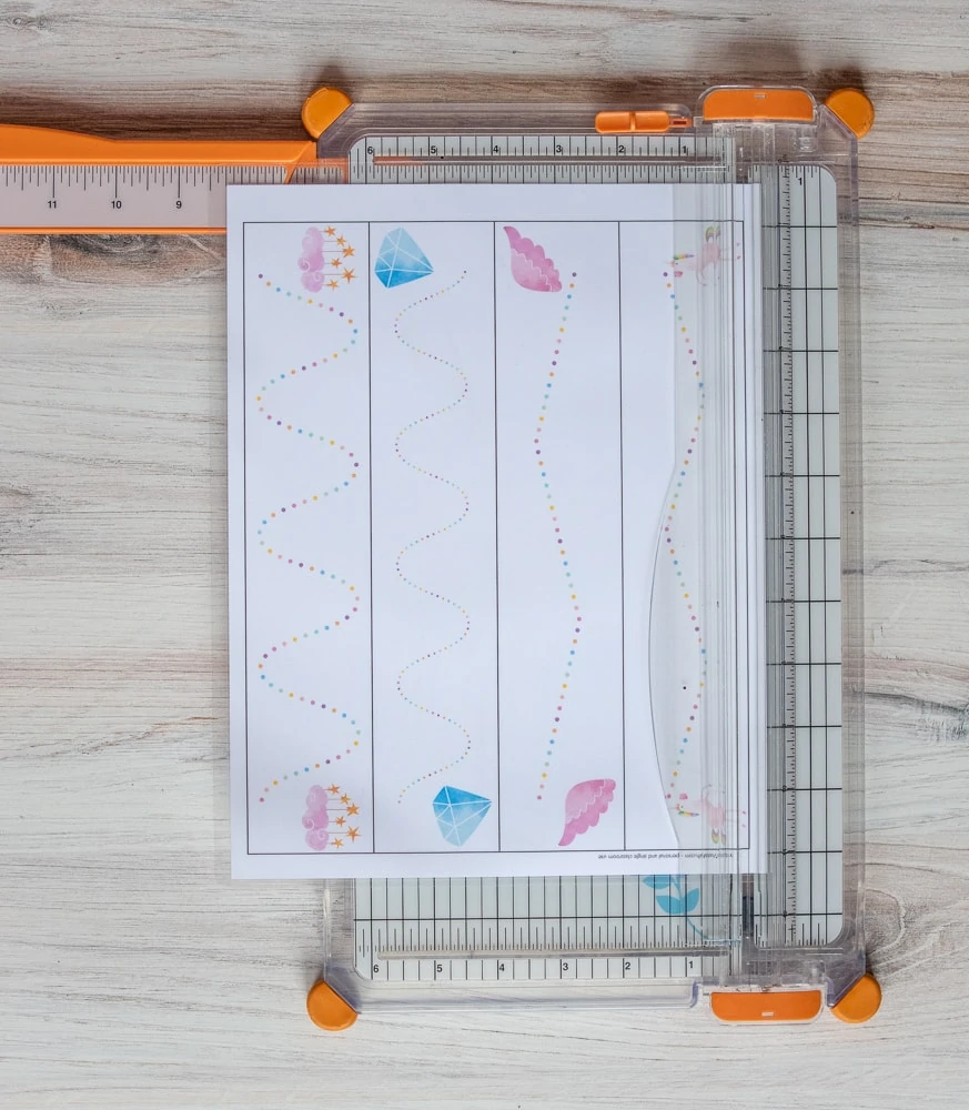 A paper trimmer on a wood surface. A printed and laminated prewriting practice page is on top of the paper trimmer. The page features four rainbow dotted paths that run the length of the page. Each line has a unicorn themed image on either end. 