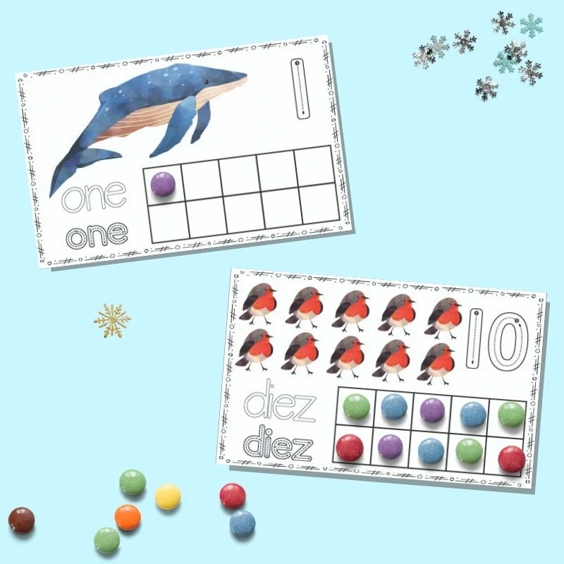 Two printable winter ten frame cards on a blue background. The upper card has "one" and a single whale with a purple candy in the first spot on the ten frame. The lower card has "diez" with ten red robins and all squares on the ten frame filled in. 