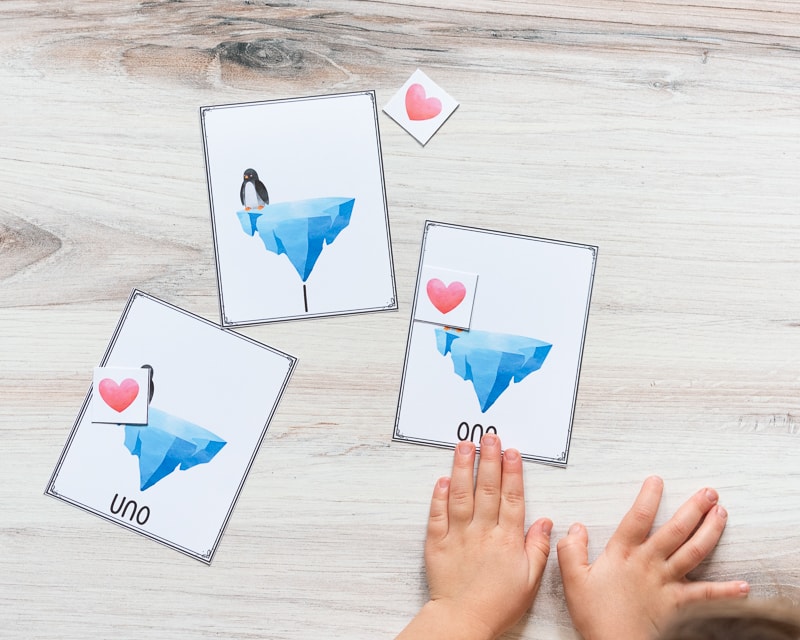 A young child's hand on a card with "one" and an iceberg on it. There is a red heart card covering a penguin on the card. There are two additional cards on the table. Both have a single card and penguin. One has "1" on the bottom and another has "uno"
