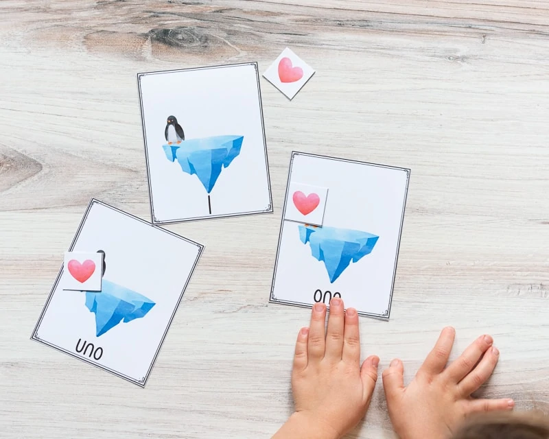 A young child's hand on a card with "one" and an iceberg on it. There is a red heart card covering a penguin on the card. There are two additional cards on the table. Both have a single card and penguin. One has "1" on the bottom and another has "uno"