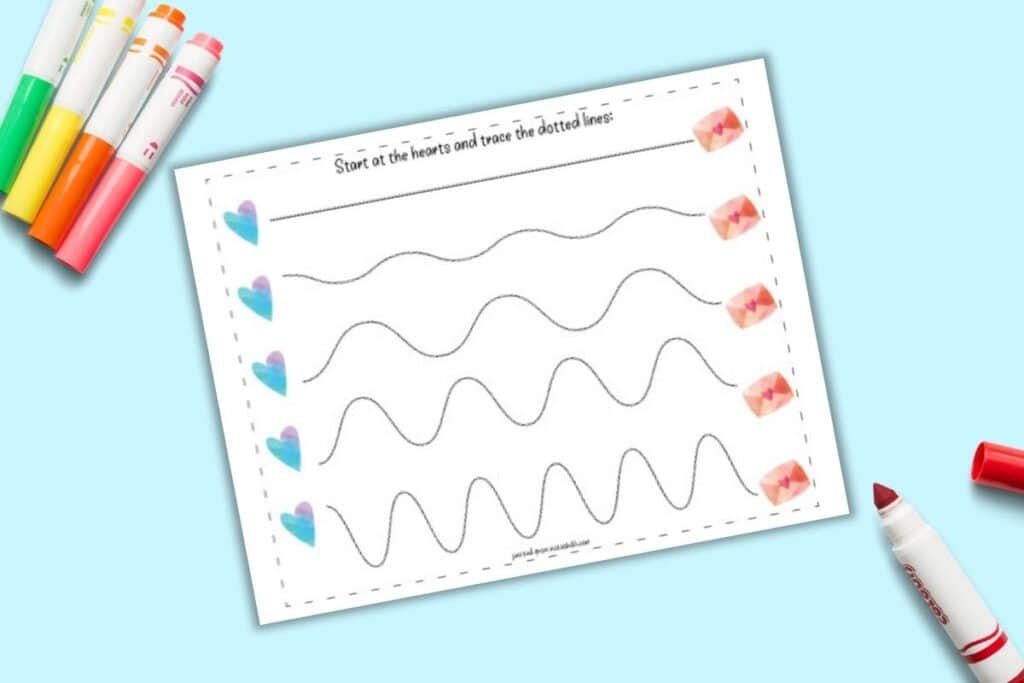 A top down flatlay mockup of a printable fine motor prewriting practice worksheet. On the left is a line of five hearts next to five wavy dotted lines. The lines lead to a Valentine's envelope. The page is on a blue background with colorful child's markers.