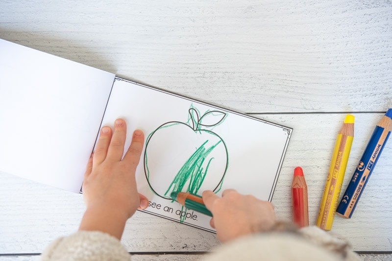 A child's hand using a green pencil to color an apple in a printable letter a book