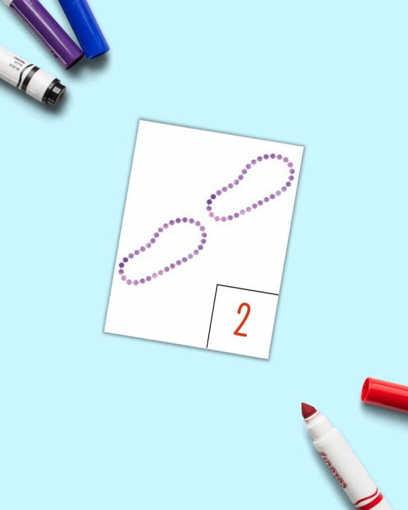 A free printable Mardi Gras count and write card with two strands of purple beads and a number "two." The card is on a blue background with colorful children's markers. 