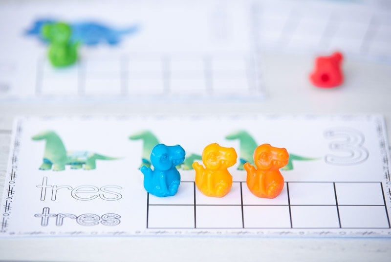 A close up photograph of a printable ten frame with three green dinosaurs and "3" and "tres." There is a ten frame with three plastic counting dinosaurs on it. 
