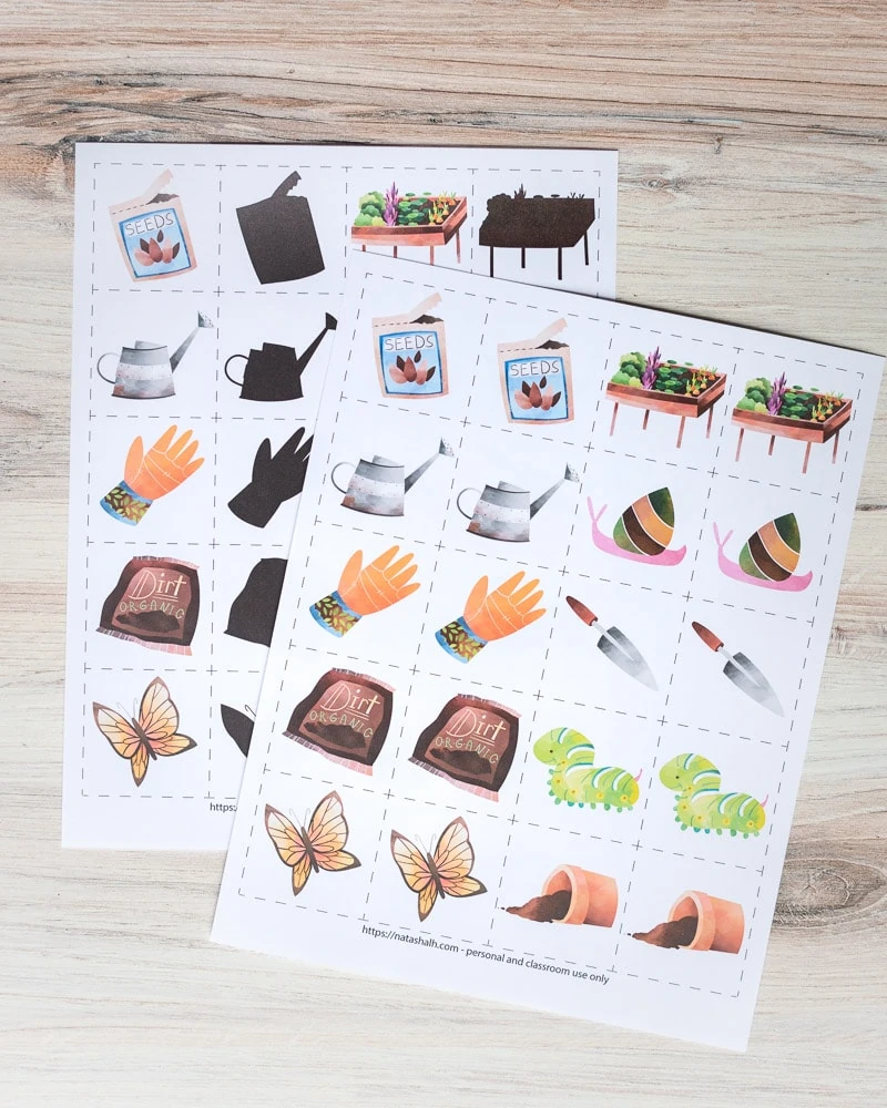 A preview of two free printable garden matching game pages. Each page has pairs of 10 garden related images inside dotted squares to cut out and use as a matching card game. The page in back has half of the images in color and the other half "blacked out" for a shadow matching game. The two printables are on a wood surface.