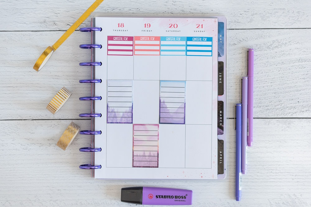 An open Happy Planner Classic with a gratitude journal sticker across the top of each column in a weekly spread page. There are also three full box note stickers. The stickers are full boxes for Happy Planner. Each box has a watercolor landscape background and a translucent white rectangle with seven lines to write on. The planner is on a white wood surface with purple pens, a purple highlighter, and gold washi tape.