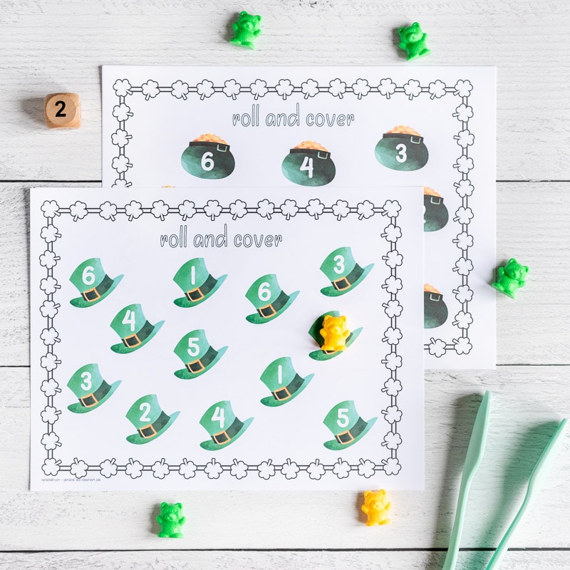 A printable St. Patrick's Day roll and cover page with 12 green top hats. Each hat has a number 1-6. One hat has a counting bear covering the number. A wood die showing "2" and a paint of tongs are next to the printable page. A second roll and cover page with pots of gold is visible behind the first.