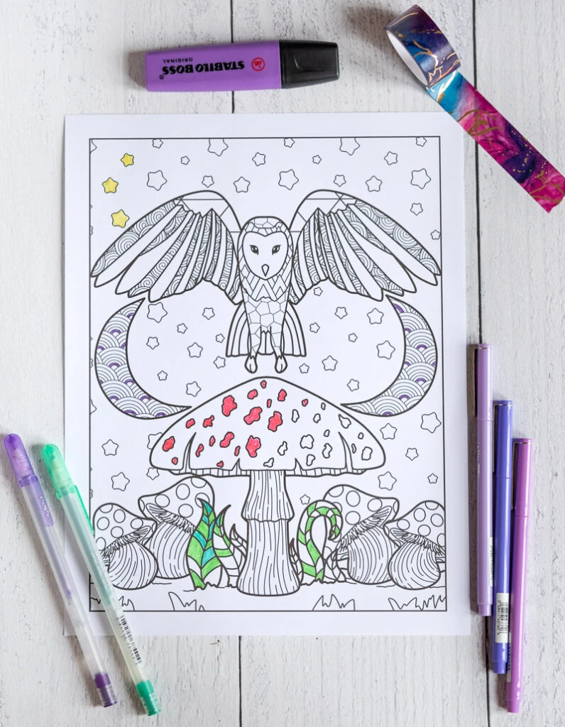 A printed owl coloring page with an owl over a mushroom with two crescent moons in the background. The page is partially colored and on a white wood surface with purple pens, a purple highlighter, and purple and blue washi tape