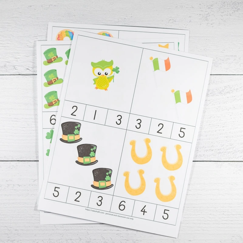 Printable St. Patrick's Day counting clip cards on a white wood surface. The font page is the only one fully visible. It has numbers 1-4 on four cards to cut out. Images include one owl, two Irish flags, three hats, and four horseshoes. 
