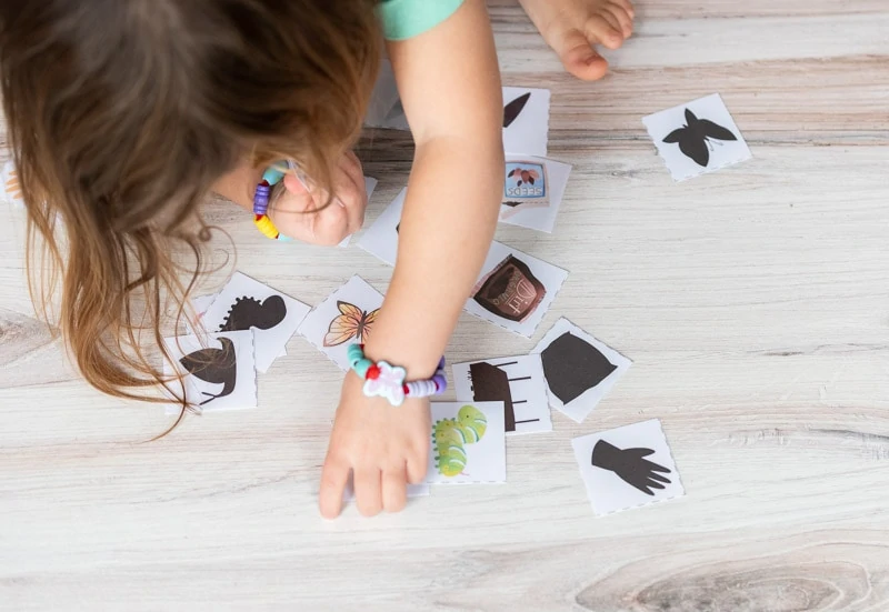 A top down view of a young girl wearing butterfly bracelets playing with shadow matching cards on a wood floor.