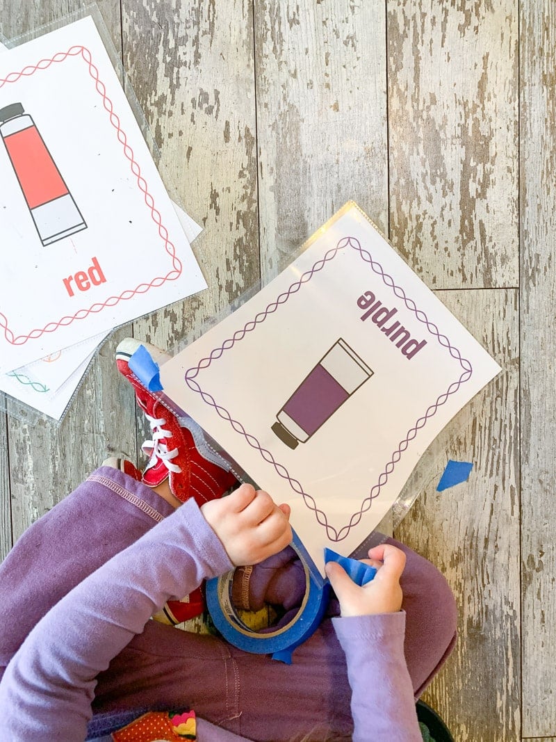 A top down view of a young child wearing purple clothes and red shoes. She is sitting on the floor placing a piece of blue painter's tape on a poster with a purple tube of paint and the word "purple"