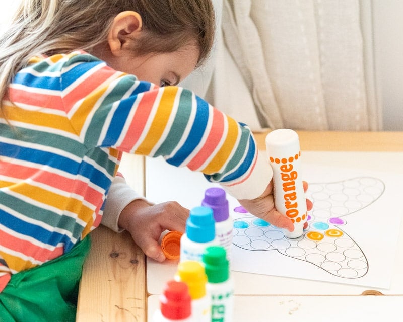 An image of a preschooler in a rainbow striped shirt using an orange dauber style maker to color a butterfly dot marker coloring page 
