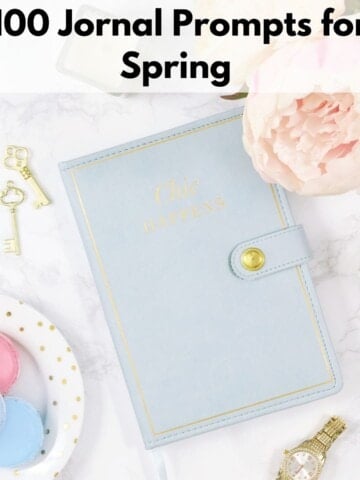text overlay "100 journal prompts for spring" over an image of a blue notebook with macrons and pink flowers on a white marble surface