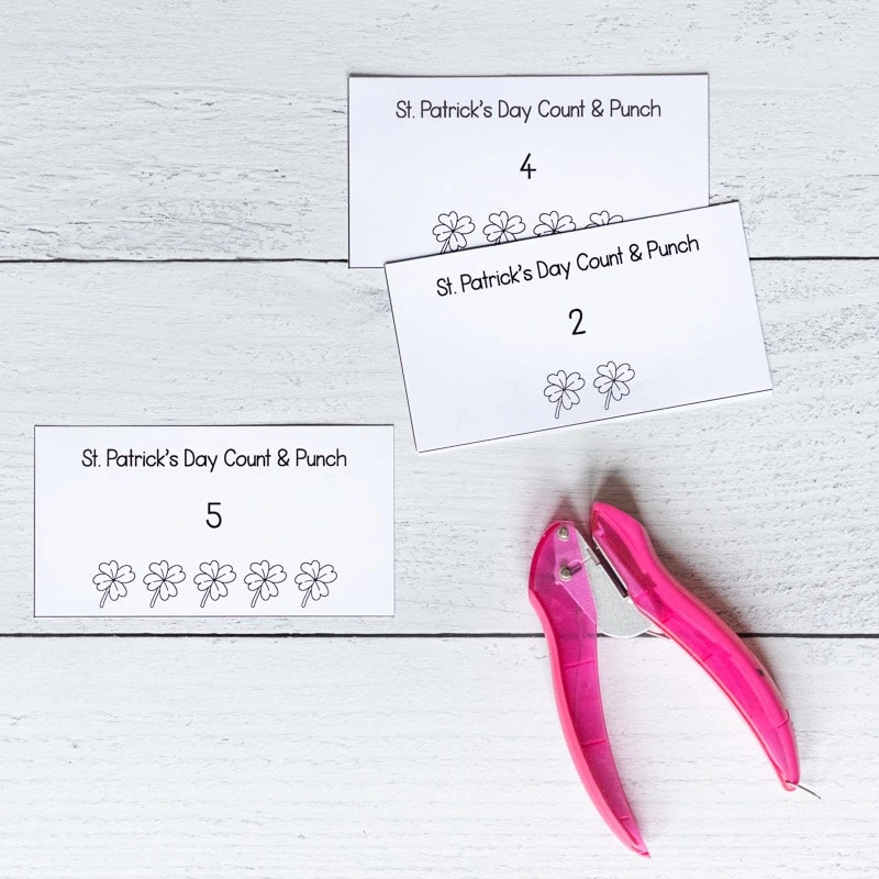 Three printed and cut St. Patrick's Day count and punch cards on a white wood surface with a pink single hole punch