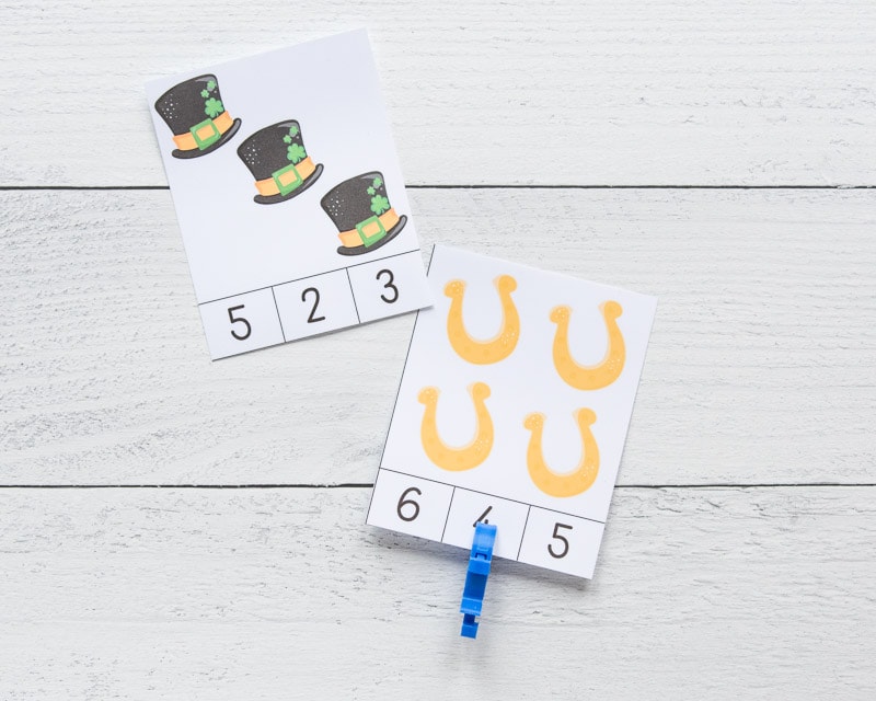 Two printed count and clip cards with a St. Patrick's Day theme. One card has four gold horseshoes and the number "4" is clipped with a blue clip. Next to this card is a card with three black hats.