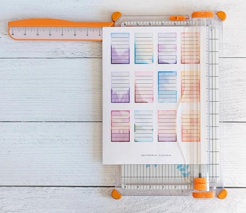 Trimming a page of printable planner stickers with a paper trimmer. The stickers are full boxes for Happy Planner. Each box has a watercolor landscape background and a translucent white rectangle with seven lines to write on.