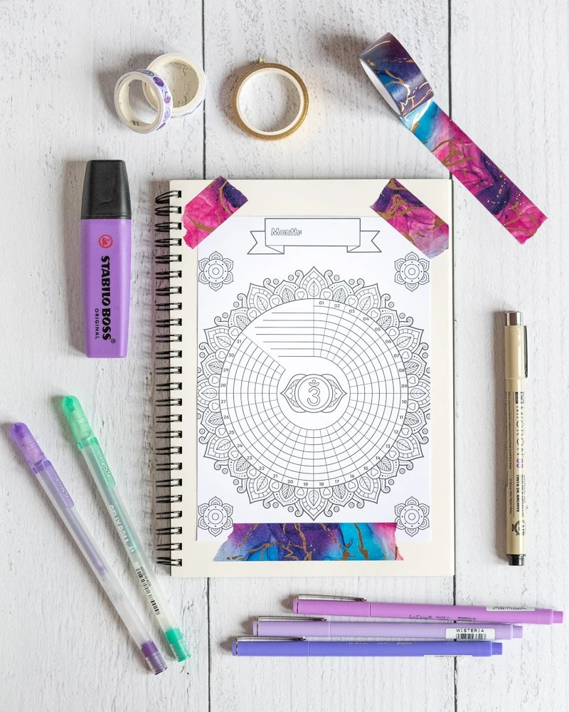 A spiral bullet journal with a charka themed bullet journal printable taped to a page with marveled puree and blue washi tape. The planner is surrounded by offie supplies, including gel pens, a purple Stabilo Boss highlighter, a Micron pens and three purple fineliners. 