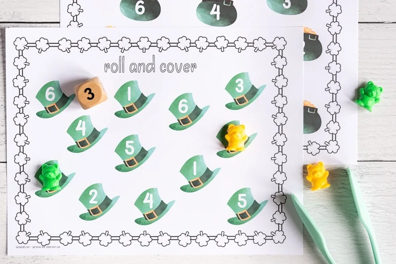 A printable St. Patrick's Day roll and cover page with 12 green top hats. Each hat has a number 1-6. Two hats have a counting bear covering the number. A wood die showing "3" and a paint of tongs are next to the printable page. 