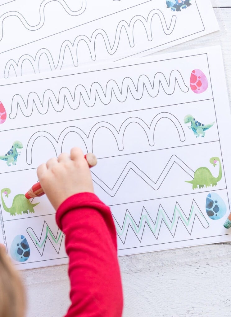 An overhead view of a young child's hard using a large wood pencil to trace a path on a dinosaur themed prewriting practice page. The page has four paths to trace inside the lines and a dinosaur on each end.