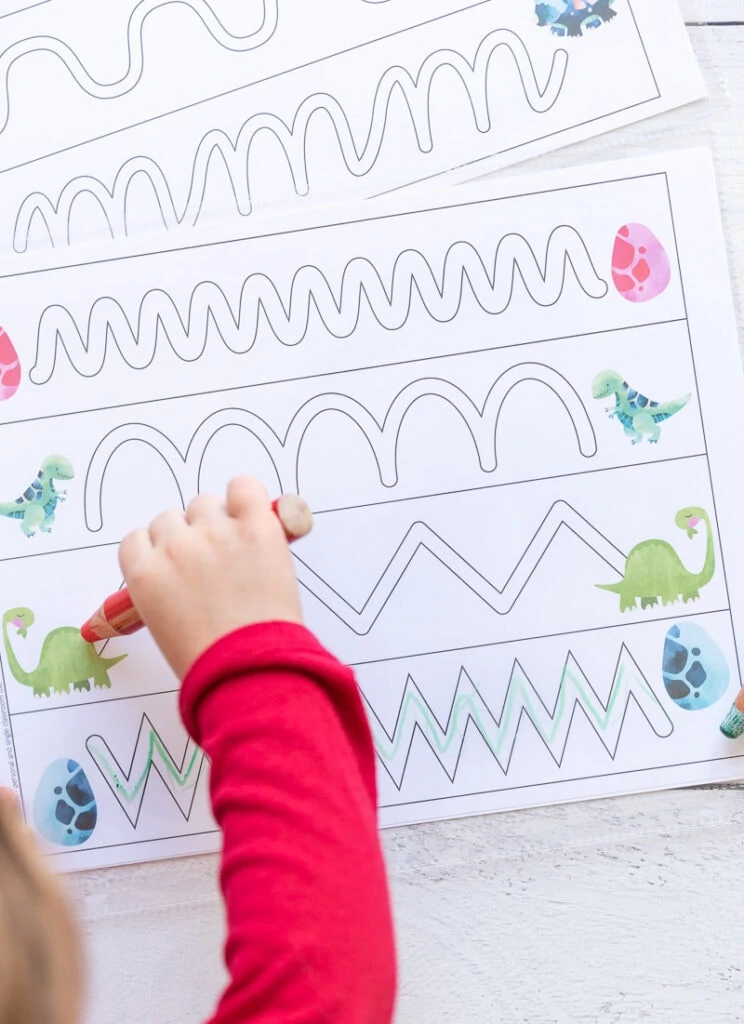 A preschooler's arm in a  red shirt holding a red crayon to trace a dinosaur themed trace in the path page.