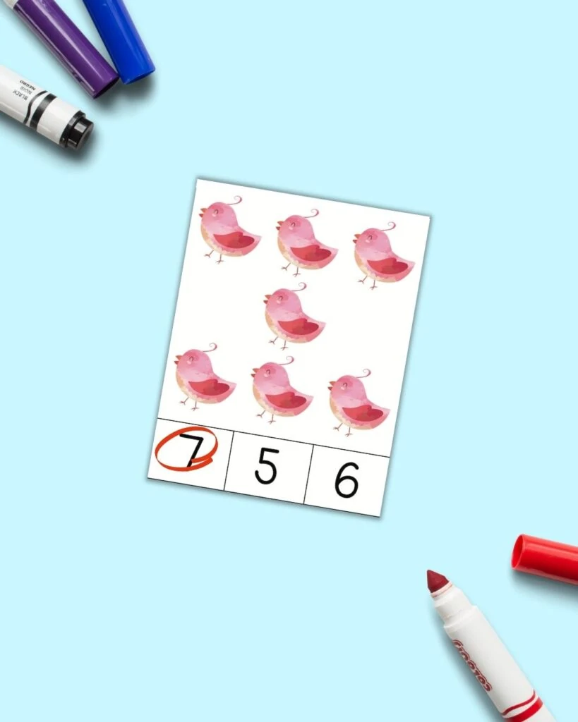 A Valetine's Day count and clip card with seven pink birds and numbers 7, 5, and 6 across the bottom. The 7 is circled in red. The card is on a blue background with colorful children's markers.