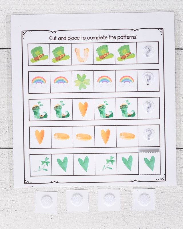 A St. Patrick's Day complete the pattern worksheet for preschoolers. Below are four square tiles turned face down. Each as a white hook and loop closure dot on it. At the end of each sequence, there is a question mark with a hook and hoop dot on top. One pattern is completed with a green heart.