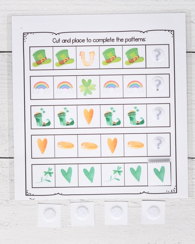 A St. Patrick's Day complete the pattern worksheet for preschoolers. Below are four square tiles turned face down. Each as a white hook and loop closure dot on it. At the end of each sequence, there is a question mark with a hook and hoop dot on top. One pattern is completed with a green heart.