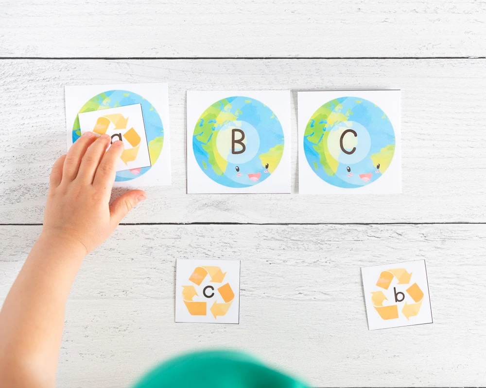 A child' s left hand placing a small printable card with a gold recycling symbol and a lowercase a on top of a larger printable card with the planet Earth and an uppercase letter A