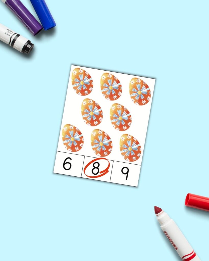 An Easter themed count and clip card with 8 orange easter eggs on it. Below are the numbers 6 8 9. The 8 is circled in red. The card is on a light blue surface with colorful children's markers. 