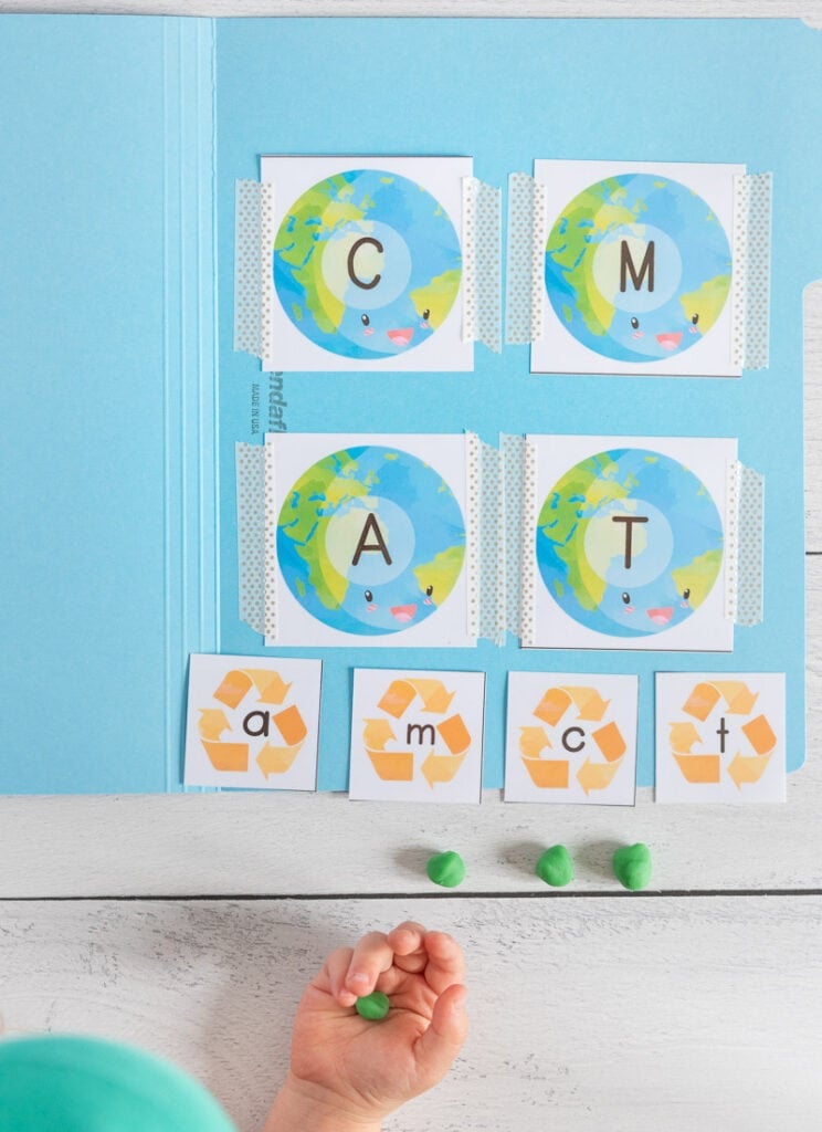 A child's hand holding a ball of green play dough. Above the hand is a blue file folder with four uppercase letter cards with the planet Earth and one letter on each card. Letters shown are C, M, A, T. Below the uppercase cards are four smaller cards with a gold recycling symbol and lowercase letters c m a t