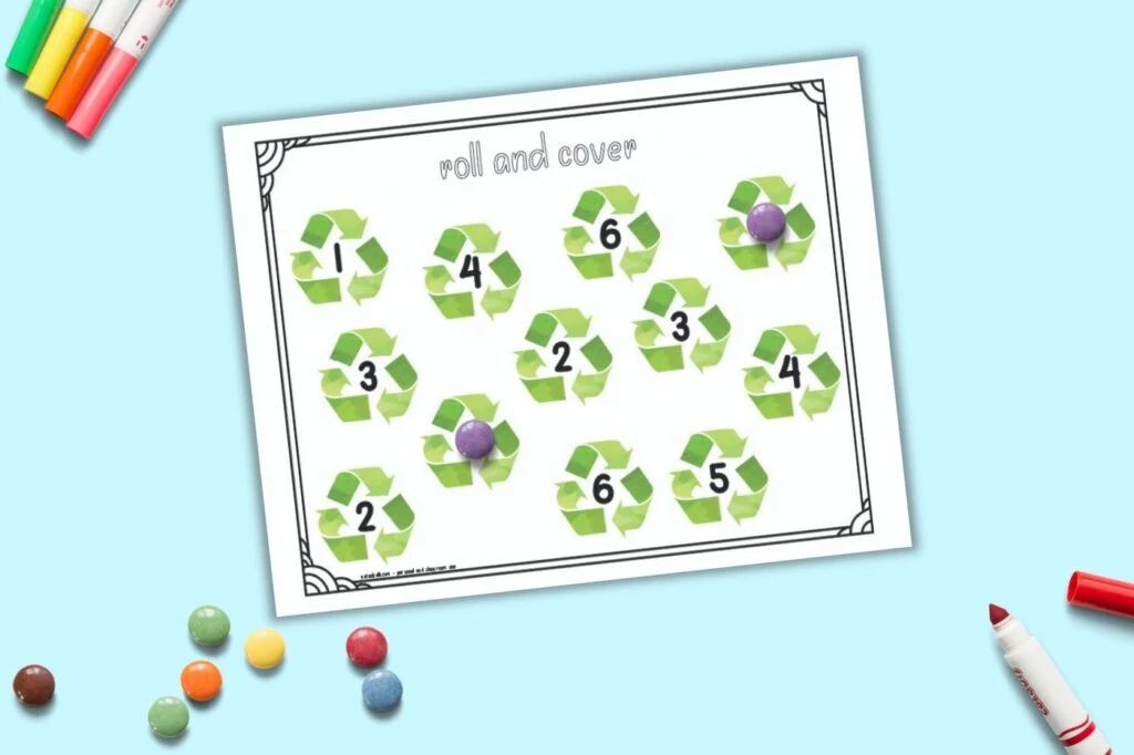 A printable roll and cover mat for preschool math. The page has the numbers 1-6 two times apiece. Each number is in the center of a recycling symbol. Purple round candies cover two of the numbers and an additional pile of candies is on the bottom left of the image.