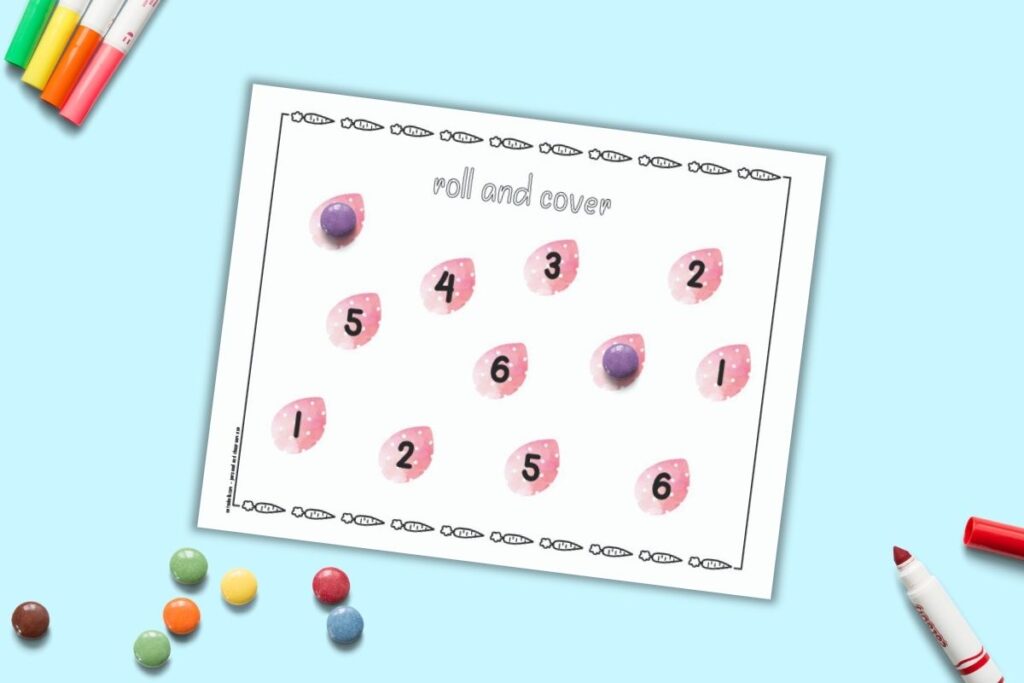 A free printable Easter roll and cover mat for preschoolers. The mat has 12 pink Easter eggs and each egg has a number 1-6 printed on it. Two numbers are covered with round purple candies. More candies are on a blue surface beside the mat.