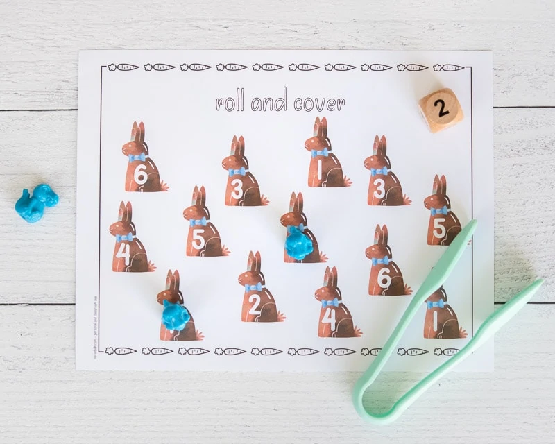 A printable Easter roll and cover mat with 12 chocolate bunny images. Each bunny clipart has a number 1-6. Two numbers are covered with small blue dinosaurs. A pair of seafoam colored plastic tongs is on the bottom right of the page.