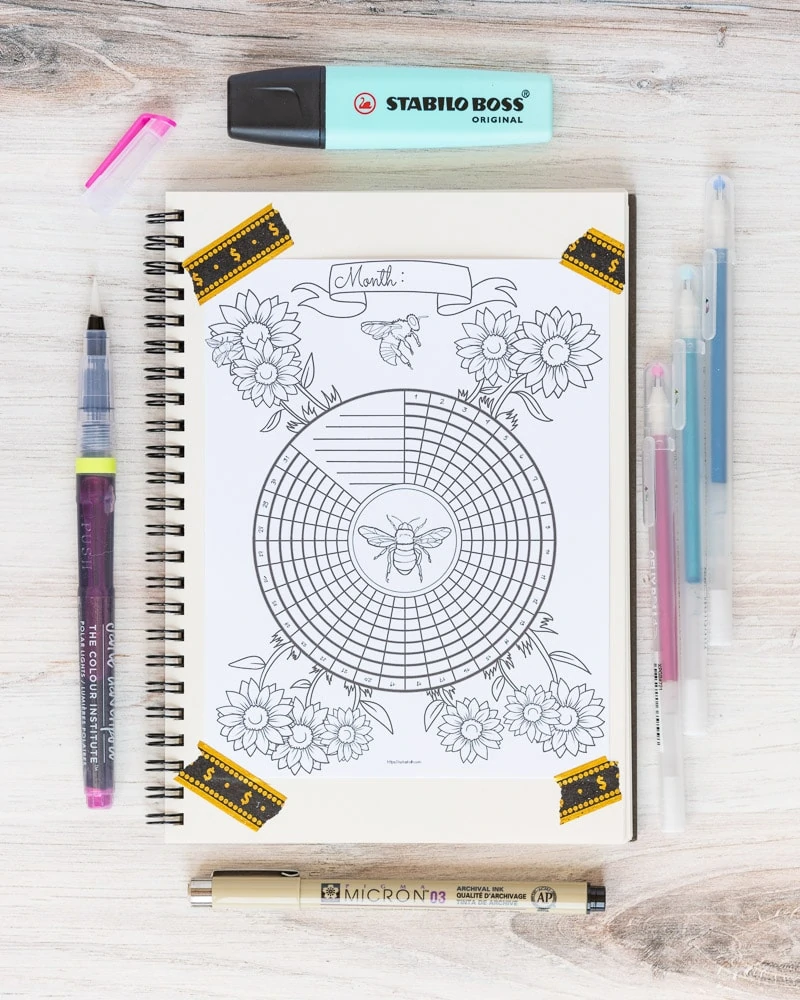 A notebook with a bee themed circular habit tracker taped to the page with washi tape. There are desk supplies around the notebook, including a brush pen, Sakura Micron pen, Stabilo highlighter, and Gelly roll pens.