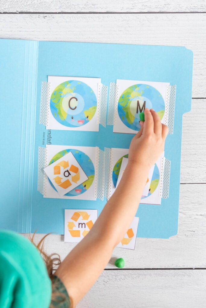 A child's hand placing a green ball of play dough on a card with an uppercase letter M on an illustrated picture of the planet Earth.