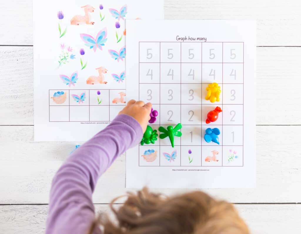 A top down shot of a preschooler using insect shaped math counters to graph results from a spring themed count and graph worksheet with numbers 1-5.