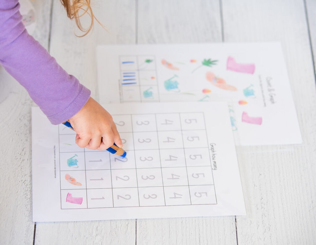 A side view shot of a preschooler wearing a purple shirt drawing on a count and graph page with a blue colored pencil.