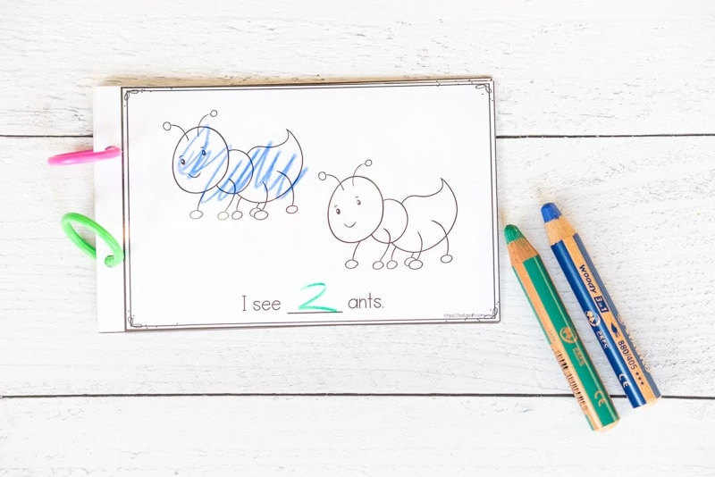 A page with two ants and the text "I see 2 ants." The two is written in green pencil and one and has been colored with blue pencil. The green and blue pencils are on the table next to the page.