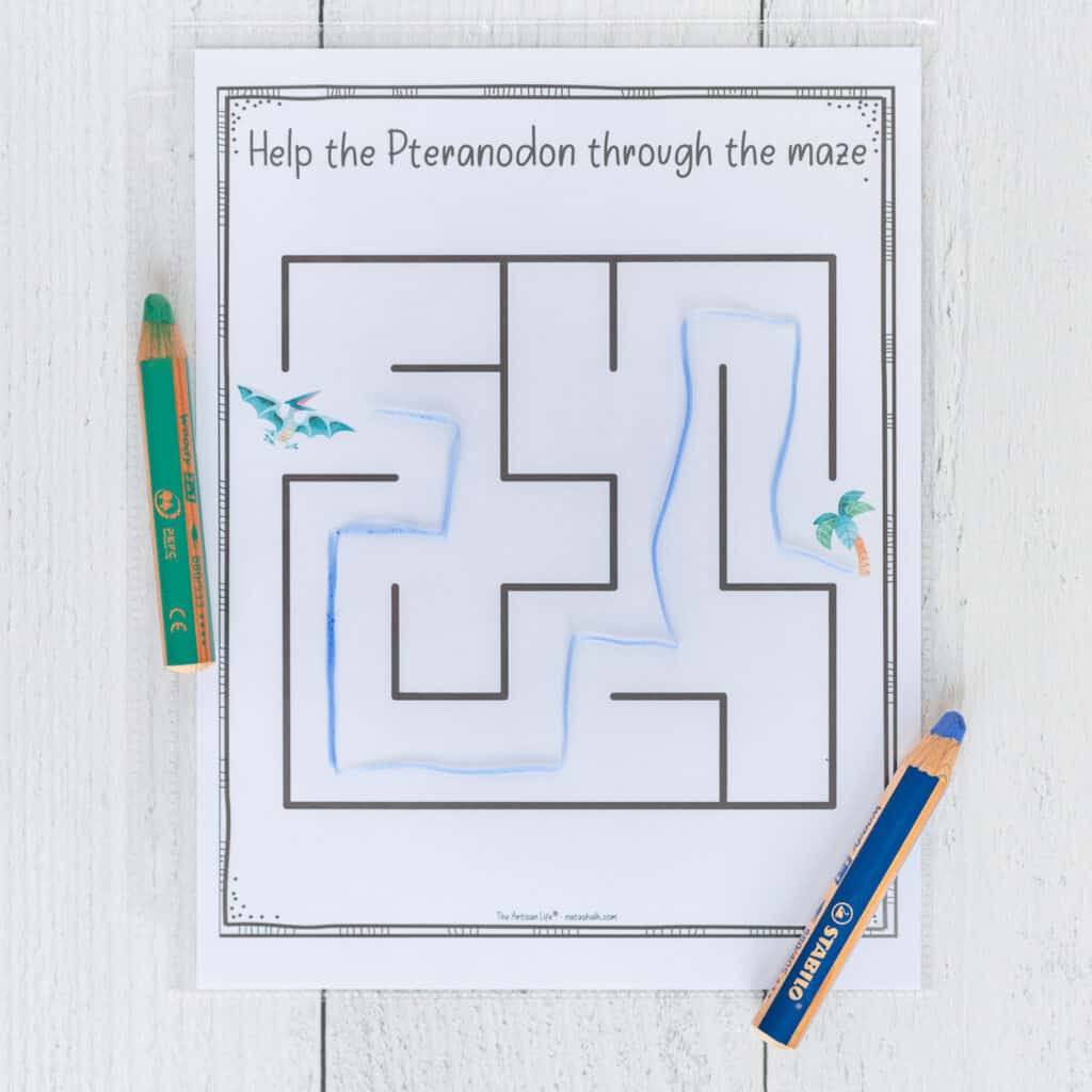 An easy maze for preschoolers featuring a pteranodon. The page is in a page protector and has been solved with a blue wax crayon. The blue wax crayon and a green wax crayon are beside the printed page.