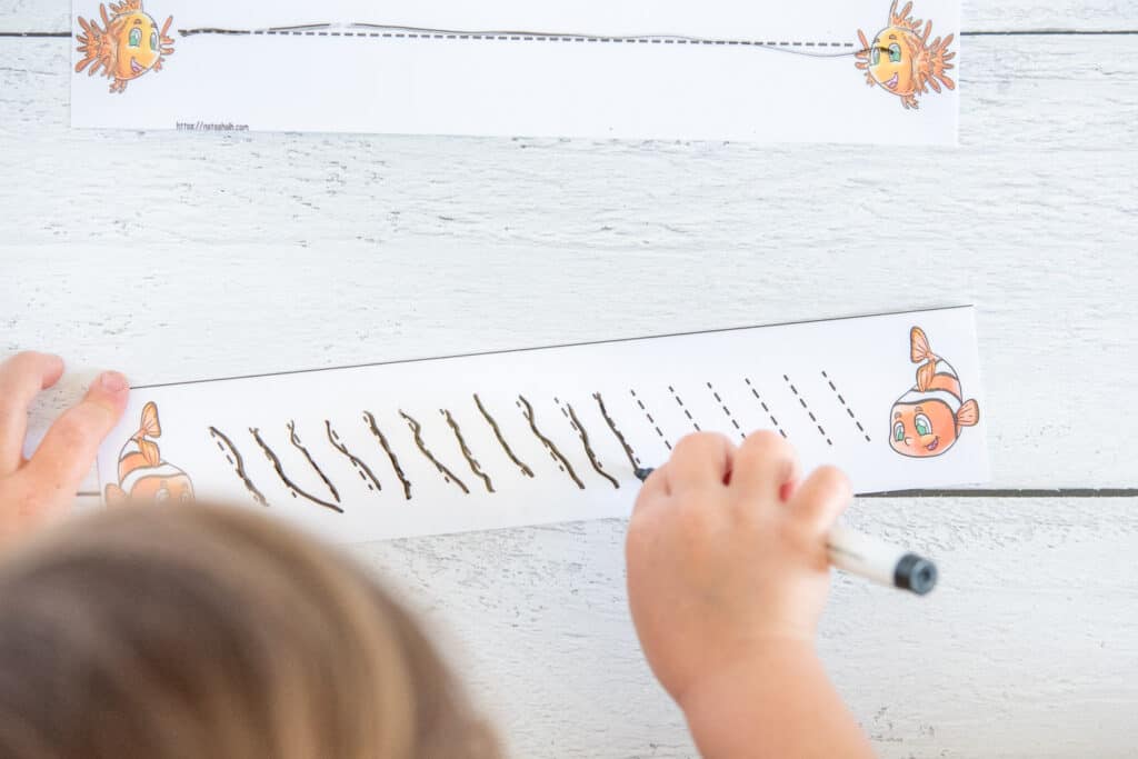 A top down view of a preschooler using a dry erase marker to trace dotted backslash marks on a laminated card. There is a clownfish on both ends of the card.
