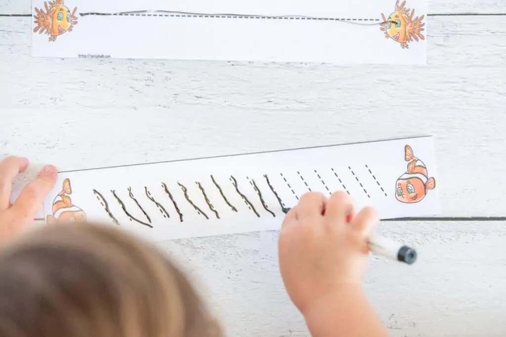 A top down view of a preschooler using a dry erase marker to trace dotted backslash marks on a laminated card. There is a clownfish on both ends of the card.