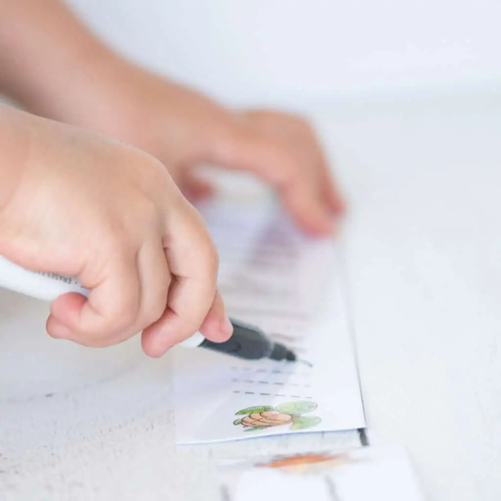 A child using a black dry erase marker to trace dotted slanted lines on a tracing card strip with a green sea turtle
